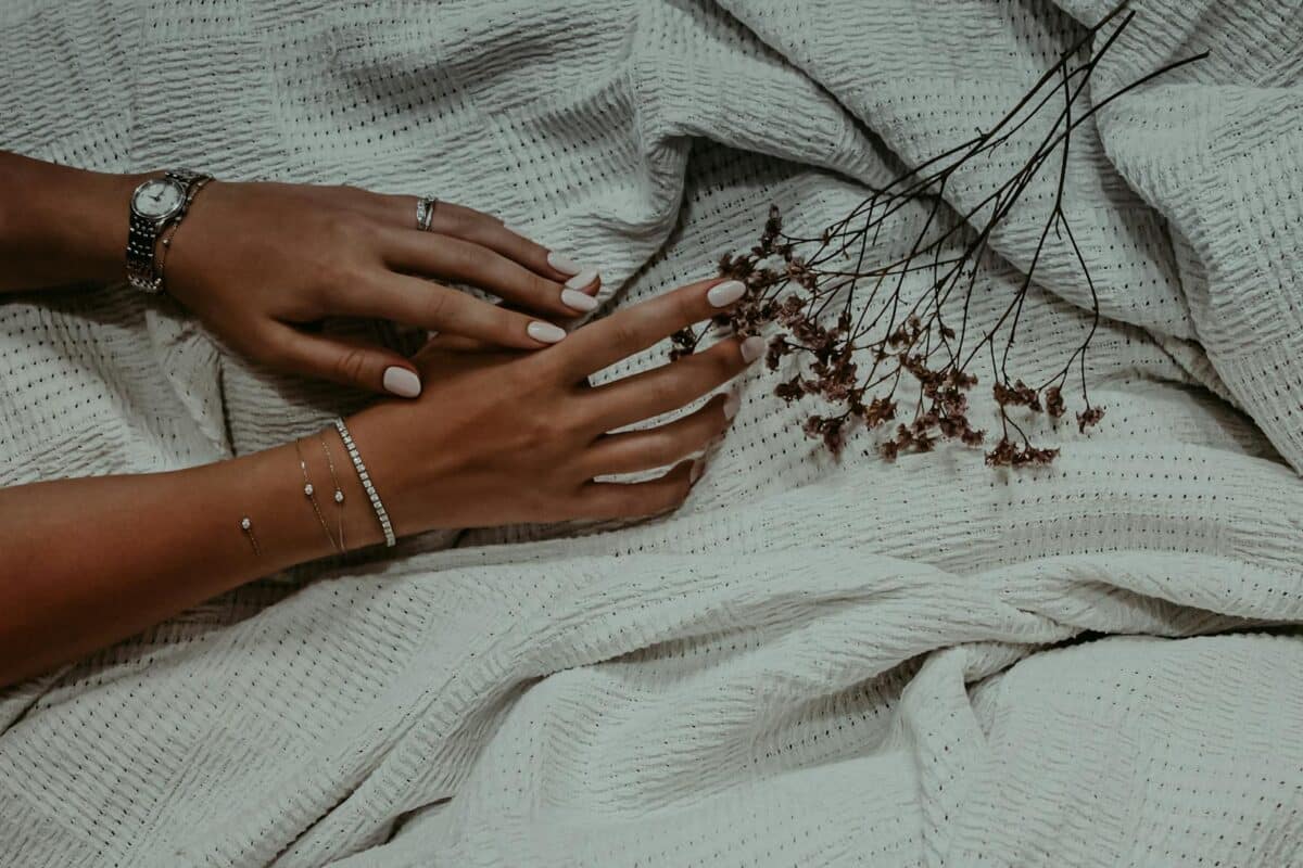 Close-up of manicured hands with elegant jewelry, resting on a textured white fabric with dried flowers.